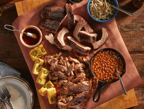 Wood cutting board topped with a sheet of butcher paper and topped with pulled pork, BBQ ribs, baked beans, cornbread and coleslaw