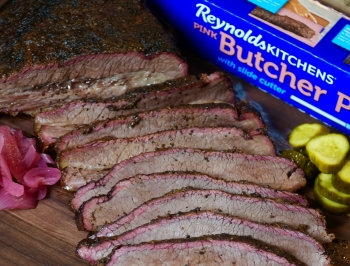 Sliced brisket sitting on a cutting board alongside a package of Reynolds Kitchens Butcher Paper